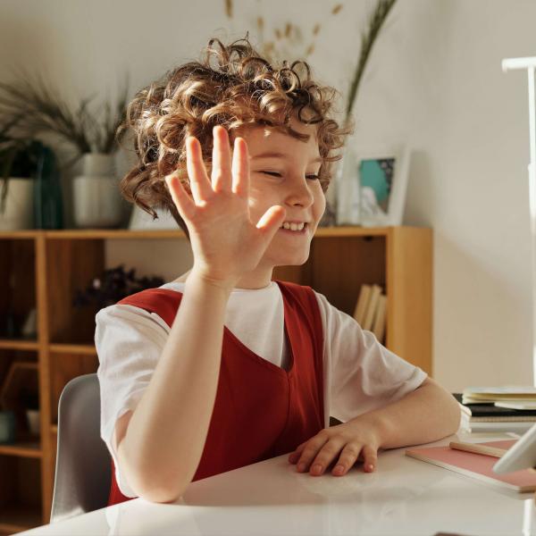 Youngster with curly red hair raises hand while looking at tablet in a bedroom