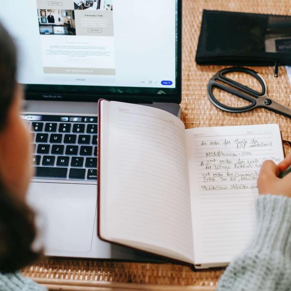 Top view of student with dark hair writing in a notebook next to a laptop on a wicker table