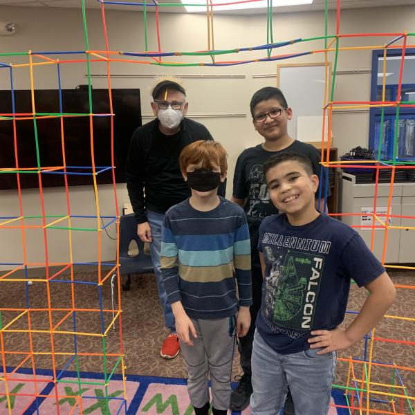 Three children and teacher standing under colorful structure inside classroom