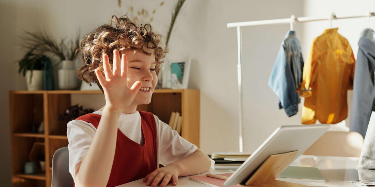 Youngster with curly red hair raises hand while looking at tablet in a bedroom