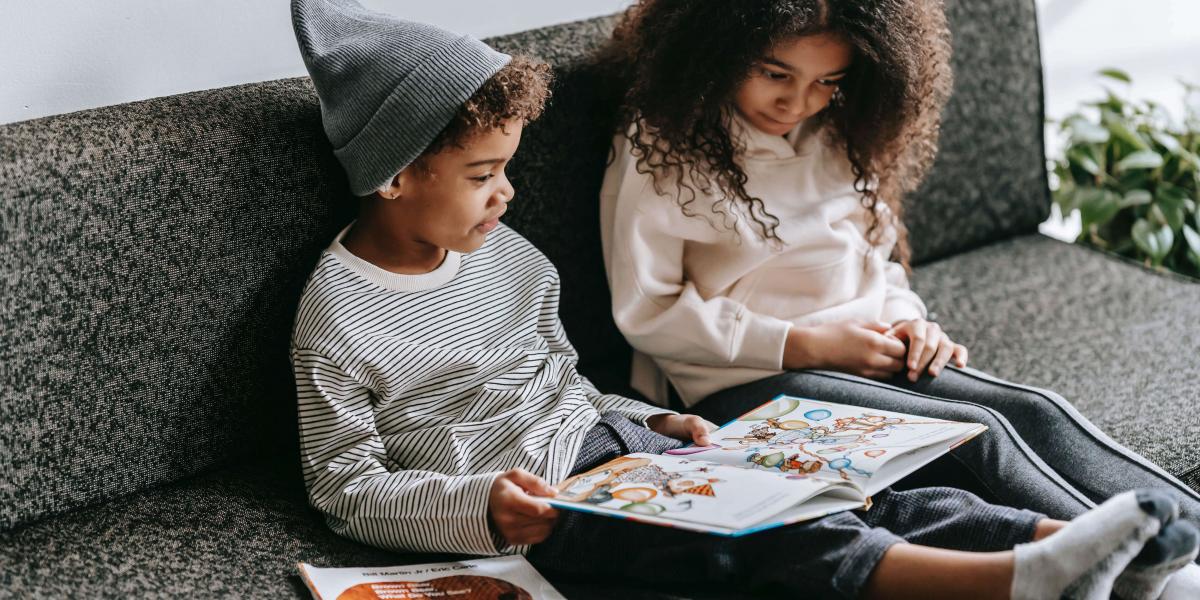 Siblings with dark hair seated on a grey couch reading to eachother