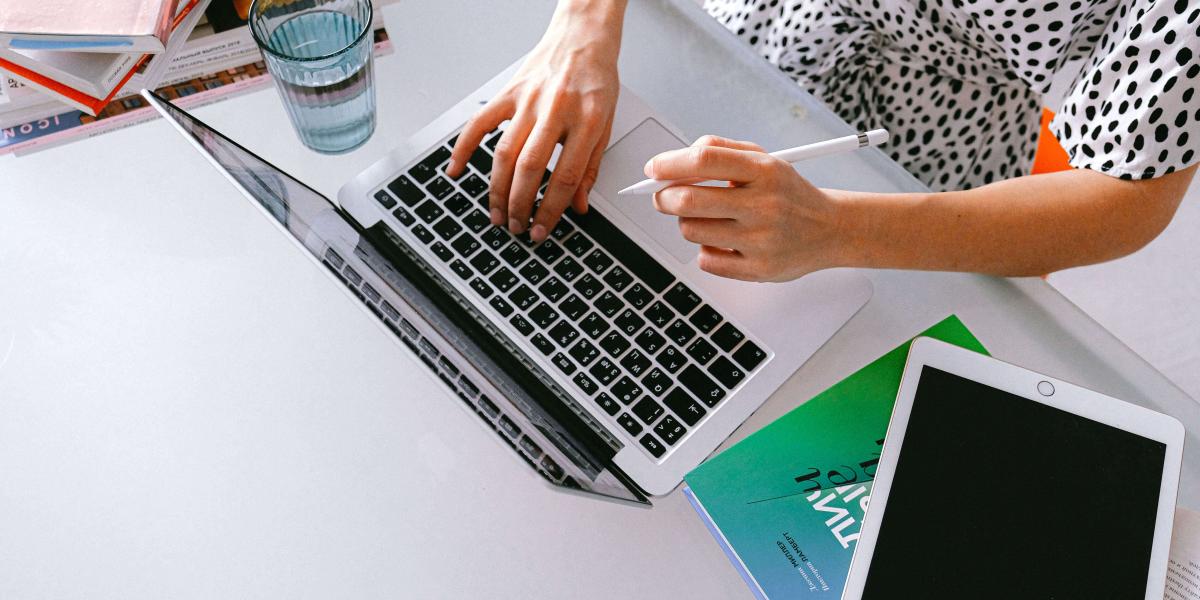 Top rear view of hands at a laptop. An iPad is lying next to the laptop