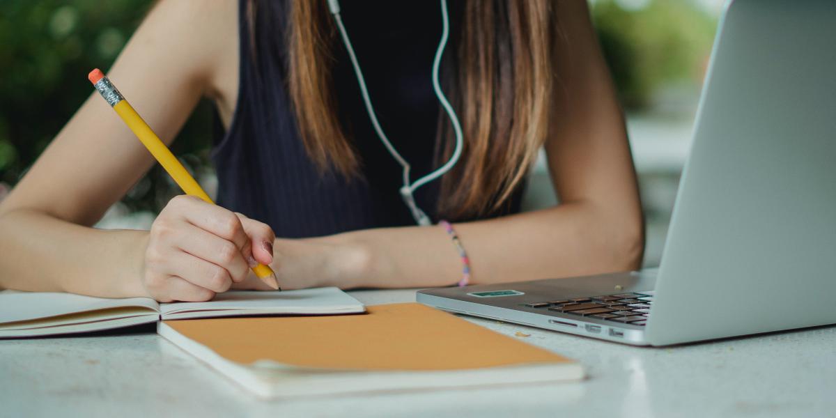 Student with long hair writing next to laptop. Head is not visible. 