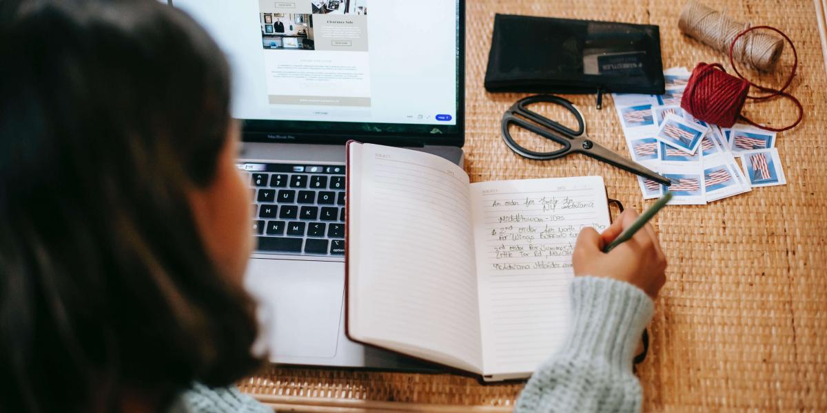 Top view of student with dark hair writing in a notebook next to a laptop on a wicker table