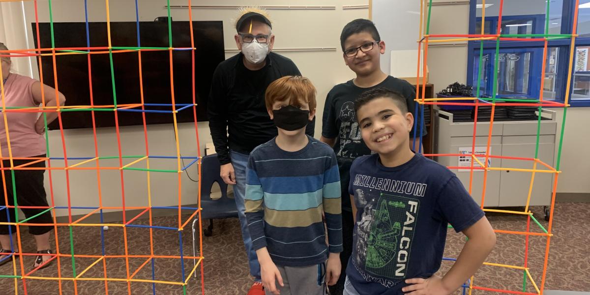 Three children and teacher standing under colorful structure inside classroom
